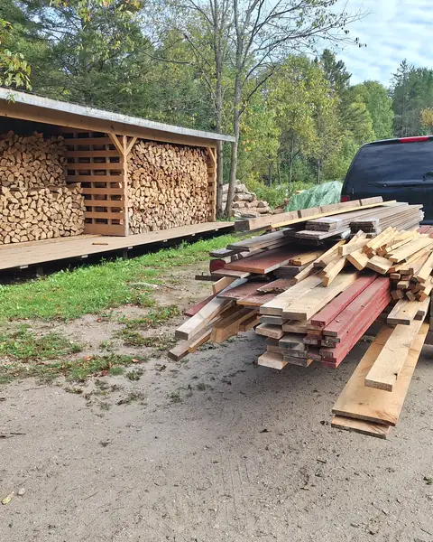 Reclaimed lumber stacked on a trailer beside a full woodshed in South River